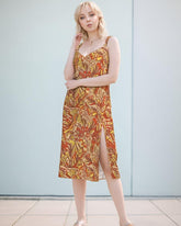 midi dress with a vibrant, psychedelic retro floral  print featuring swirling patterns in shades of red, yellow, and brown. The dress has a V-neckline, spaghetti straps, and a leg-baring side slit. Blissfully Brand